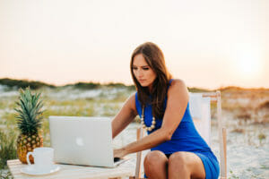 Woman with brown hair wearing blue dress works on laptop at beach